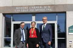 Two men and one woman stand in front of a building with a sign reading Robert Larner M.D. College of Medicine. The woman wears a medallion on a green and gold ribbon.