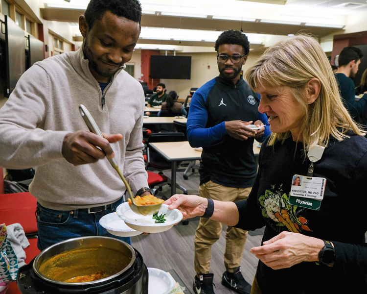 In a classroom, a person scoops orange-colored soup into a bowl held by another person, as a third person observes.
