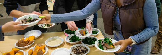 close up of several people's arms and hands passing each other bowls filled with orange sweet potatoes, green kale and black beans