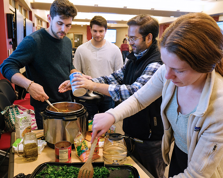 Four people cooking together in a classroom, stirring and seasoning soup in a crockpot and sauteeing kale on an electric griddle