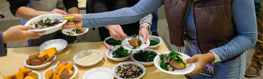 Several people's hands holding and passing paper plates and bowls containing baked sweet potatoes, hummus, black beans, kale
